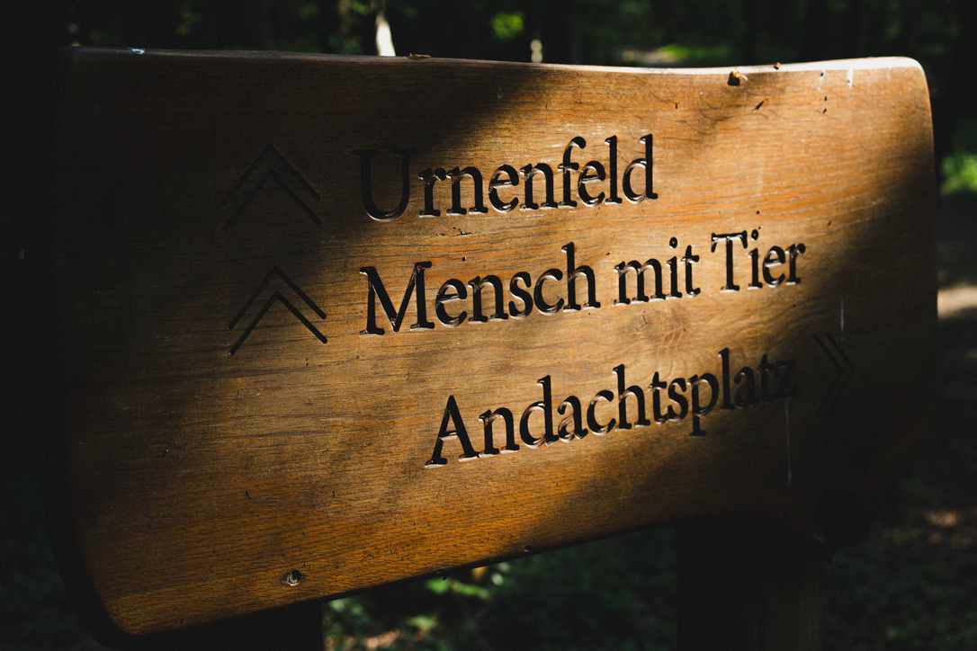 Holzschild mit Wegweisern zu Urnenfeld, Mensch mit Tier und Andachtsplatz